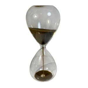 May include: Transparent glass hourglass filled with dark brown sand. The hourglass features a rounded top and bottom, joined by a slender neck. The sand is in the process of flowing from the upper to the lower chamber. A decorative item.