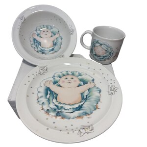 Può includere: Un set di piatti per bambini in ceramica bianca con un disegno di bambino. Il set comprende una ciotola, un piatto e una tazza. Il bambino è circondato da un disegno blu a forma di cavolo. Il piatto e la ciotola hanno piccoli puntini blu e illustrazioni di coniglietti.