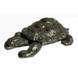 May include: A silver-toned metal turtle figurine featuring a textured, patterned shell and detailed skin on the head and limbs. The object is depicted in a crawling position with its head extended forward and legs positioned outward to the sides.