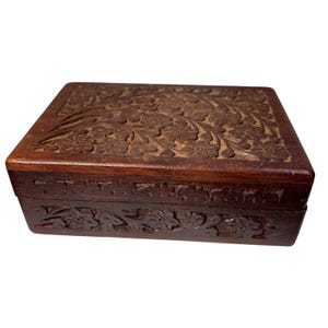 May include: A rectangular dark brown wooden box featuring intricate floral carvings on the lid and the front side. The box has a hinged lid design with a smooth finish on the edges and detailed relief patterns of flowers and leaves throughout.