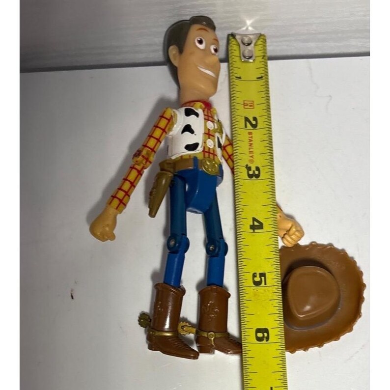 Puede incluir: Una figura de acci&oacute;n de Woody de la franquicia Toy Story. La figura lleva una camisa a cuadros amarilla y roja, un chaleco blanco, pantalones azules y botas marrones. Un sombrero de vaquero marr&oacute;n est&aacute; junto a la figura. La figura mide aproximadamente 15 cm de altura.