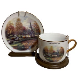 May include: A white porcelain teacup and saucer set with a gold trim. The cup and saucer feature a painting of a small church in a woodland setting. The cup has a gold chain attached to the handle and the saucer has a wooden stand. The text "Find comfort in faith" is printed on the cup.