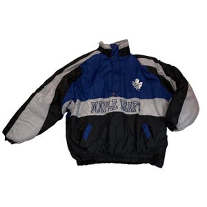 May include: A blue, black, and grey jacket with the words "MAPLE LEAFS" across the chest. The jacket has a white maple leaf logo and a high collar. The jacket is a size large.