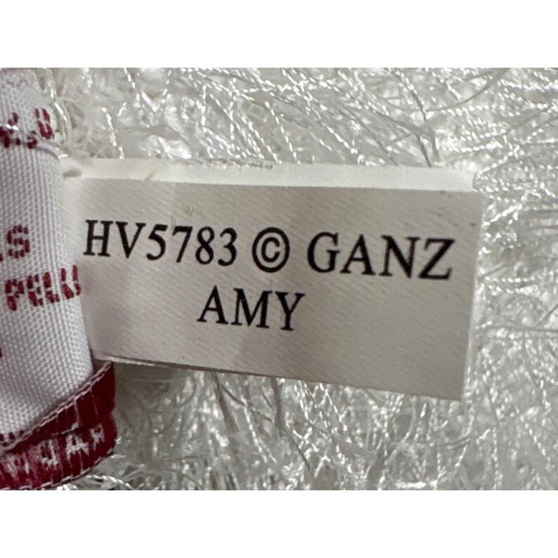 Op de afbeelding: Een wit label met de tekst "HV5783 &copy; GANZ AMY" in het zwart gedrukt. Het label is gedeeltelijk zichtbaar tegen een witte, pluizige achtergrond. De rand van een rood-witte stof is links zichtbaar.