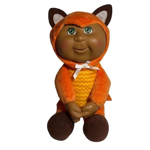 May include: A plush Cabbage Patch Kid doll wearing an orange and yellow fox costume. The doll has brown skin, green eyes, and a big smile.