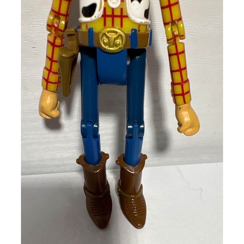 Puede incluir: Figura de acci&oacute;n de Woody, con camisa amarilla y mangas de cuadros rojos, pantalones azules y botas marrones. La figura tiene una funda marr&oacute;n y una hebilla dorada. Los brazos y las piernas de la figura son articulados.