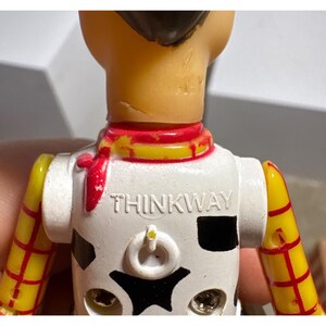 Puede incluir: Primer plano de una figura de acci&oacute;n vintage de Woody. La figura tiene un torso blanco con un patr&oacute;n de piel de vaca negra y la palabra "THINKWAY" impresa en el pecho. Presenta un pa&ntilde;uelo rojo, brazos amarillos con rayas rojas y un sombrero marr&oacute;n.
