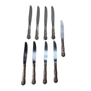 Puede incluir: Una colección de nueve cuchillos antiguos de plata. Los cuchillos tienen un diseño floral decorativo en los mangos y una hoja curvada. Las hojas son plateadas y los mangos son de un color plateado más oscuro.