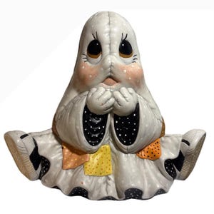 Könnte beinhalten: Eine Geisterfigur aus Keramik mit einem weißen, lakenartigen Körper, schwarz-weißen Punkten, großen dunklen Augen und rosigen Wangen. Der Geist trägt einen Rock im Patchwork-Stil mit orangefarbenen und gelben Quadraten und sitzt mit ausgestreckten Beinen.