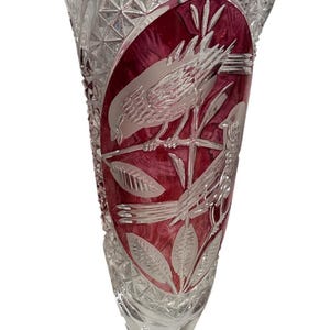 May include: A clear crystal vase with a flared top and a round base. The vase features a ruby red oval design with etched birds and leaves. The crystal is cut with geometric patterns and a scalloped rim. A decorative piece for the home.