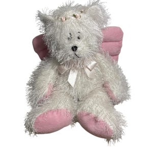 Op de afbeelding: Een witte teddybeer met roze accenten, waaronder de poten en vleugels. De beer heeft een kleine bloemenkrans en een witte strik. De vacht van de beer is harig en zacht, waardoor hij er knuffelig uitziet.
