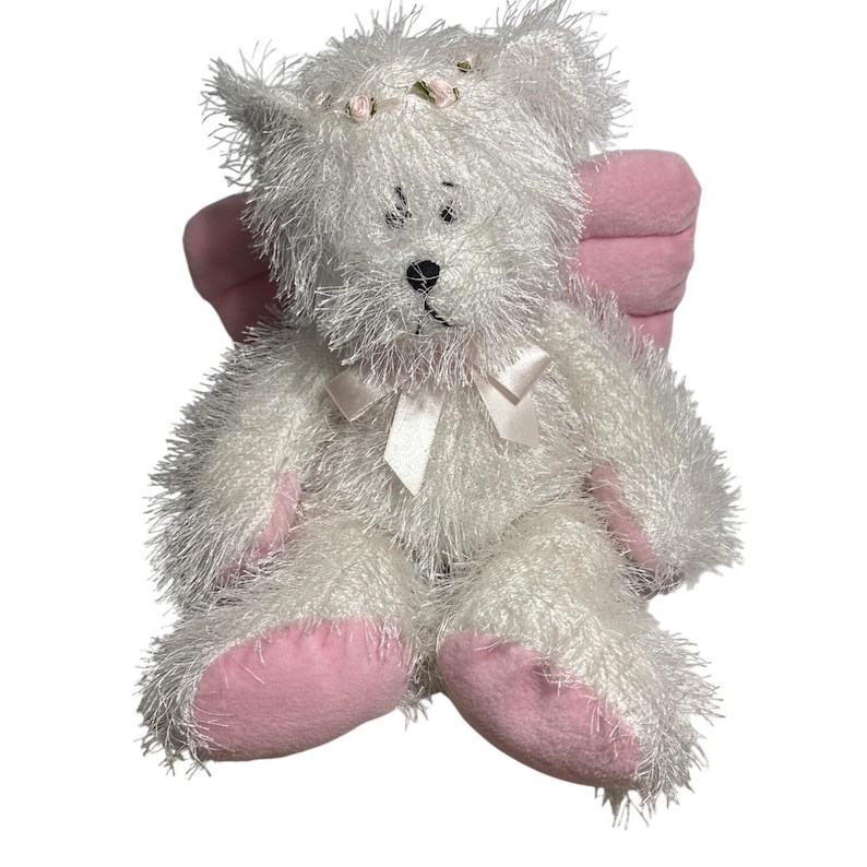 Op de afbeelding: Een witte teddybeer met roze accenten, waaronder de poten en vleugels. De beer heeft een kleine bloemenkrans en een witte strik. De vacht van de beer is harig en zacht, waardoor hij er knuffelig uitziet.