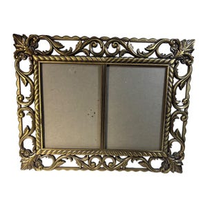 May include: A rectangular double photo frame with an antique brass finish. The frame features an ornate border with intricate scrollwork, leaf motifs, and a twisted rope pattern surrounding two side-by-side rectangular openings for photographs.