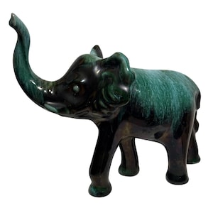 May include: A ceramic elephant figurine with a raised trunk. The elephant is glazed in shades of green and black, with a glossy finish. The figurine has a detailed design and is standing on four legs.