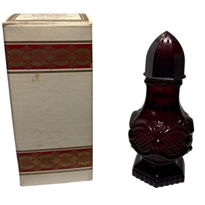 May include: A vintage, dark red glass perfume bottle with a decorative design, next to its original box. The bottle has a unique shape with a pointed top and ornate detailing. The box is cream-colored with red and gold accents.