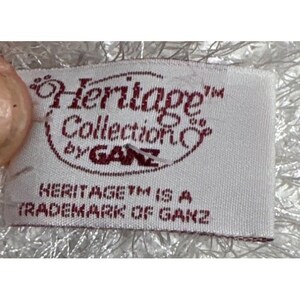 Op de afbeelding: Een close-up van een wit stoffen label met de woorden "Heritage Collection by GANZ" in kastanjebruin schrift. De tekst "HERITAGE IS A TRADEMARK OF GANZ" is hieronder afgedrukt. Het label wordt gedeeltelijk bedekt door een licht, pluizig materiaal.