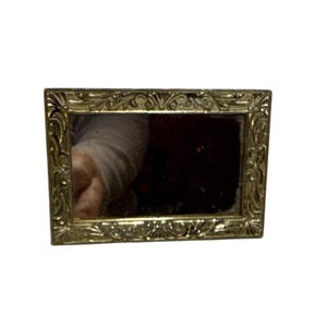 May include: A small, rectangular mirror with a gold-coloured, ornate frame. The frame has a detailed, floral-like design. The mirror's surface reflects a dark, blurred image. The overall aesthetic suggests a vintage or antique style.