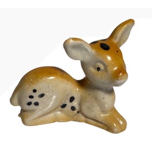 May include: A small ceramic figurine of a fawn in a resting position. The fawn is light tan with a glossy finish, featuring dark brown spots on its back and head, large ears, and a smooth, rounded body shape with a gentle, alert expression.