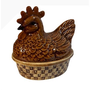 May include: A brown ceramic chicken-shaped lid sits atop a round base designed to resemble a woven basket. The chicken features detailed moulded feathers, a prominent comb, and a wattle, while the base has a textured, cross-hatched pattern.