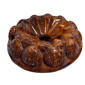 May include: A brown, ceramic, decorative object with a central hole and a scalloped edge. The object is adorned with seashell-like details around the perimeter. The glossy finish enhances the intricate design.