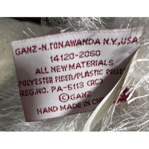 Op de afbeelding: Close-up van een rechthoekig rood label met witte tekst. Het label leest "GANZ-N.TONAWANDA N.Y., USA", "ALL NEW MATERIALS" en "HAND MADE IN CH". Het label is bevestigd aan een wit, pluizig materiaal.