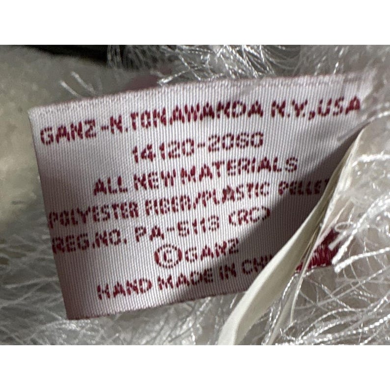 Op de afbeelding: Close-up van een rechthoekig rood label met witte tekst. Het label leest "GANZ-N.TONAWANDA N.Y., USA", "ALL NEW MATERIALS" en "HAND MADE IN CH". Het label is bevestigd aan een wit, pluizig materiaal.