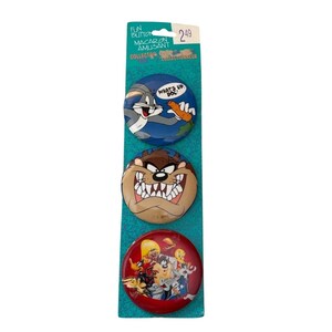 May include: A set of three cartoon character buttons on a teal backing. The top button features Bugs Bunny with the text "WHAT'S UP DOC?". The middle button shows Taz, and the bottom button has a group of Looney Tunes characters.