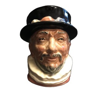 May include: A ceramic mug shaped like a man's head wearing a black top hat and a white ruff collar. The mug has a brown and white painted face with a moustache and beard.