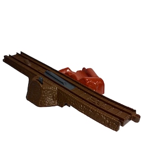 May include: A brown plastic toy train track with a rocky base and a red plastic mountain feature. The track has two parallel rails and a grey section in the middle. The toy is designed for children's play.