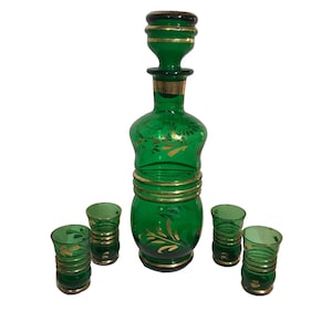 Può includere: Un set di caraffa in vetro verde con tappo abbinato e quattro bicchierini. Il set presenta dettagli dorati e motivi floreali. La caraffa e i bicchieri hanno fasce dorate.