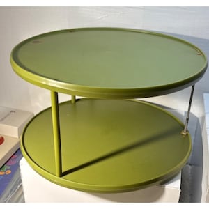 May include: A two-tiered, round, olive-green serving tray. The top and bottom tiers are connected by three vertical supports and a single metal support. The tray is made of a solid material and has a simple, functional design.
