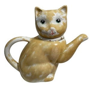 Op de afbeelding: Een keramische theepot in de vorm van een kat, in een beige en witte kleurstelling. De kat heeft groene ogen, een witte kraag en een tuit die zich uitstrekt vanaf zijn arm. De theepot is ontworpen om op een zittende kat te lijken.