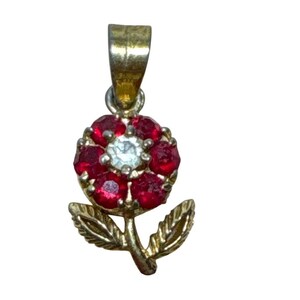 May include: A gold-toned pendant in the shape of a flower. The flower has red gemstone petals surrounding a central clear gemstone. The pendant includes a stem and leaves, all in a gold tone. A loop is at the top for a chain.