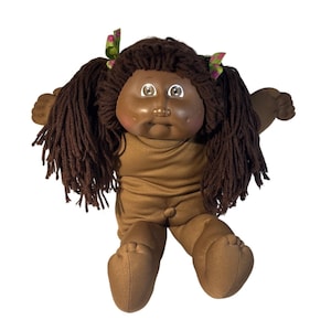 May include: A vintage Cabbage Patch Kid doll with dark brown hair in pigtails, decorated with green and pink floral ribbons. The doll has brown skin, a brown onesie, and outstretched arms.