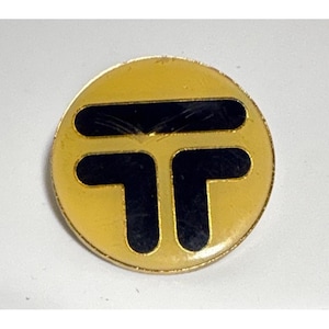May include: A round, gold-colored pin with a black, stylized letter "T" design. The pin has a shiny finish and appears to be made of metal. The design is simple and geometric, with a bold, graphic look.