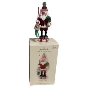 Puede incluir: Adorno navideño Hallmark Keepsake Candy Claus. La figura de Papá Noel viste un abrigo rojo, botas negras y barba blanca. Sostiene un calcetín verde y un pequeño árbol de Navidad. El adorno está sobre una base a rayas rojas y blancas.