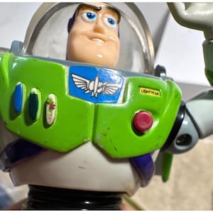 May include: Close-up of a Buzz Lightyear action figure. The toy has a green and white spacesuit with blue and red accents. The face is a light skin tone with blue eyes and a smiling expression. The word "LIGHTYEAR" is visible.