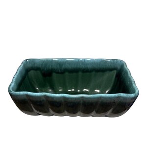 Puede incluir: Una jardinera rectangular de cerámica color turquesa con un diseño festoneado. La jardinera tiene un acabado brillante y un interior verde oscuro. Los bordes de la jardinera son de un tono turquesa más claro.