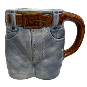 May include: A ceramic mug designed to resemble a pair of blue jeans. The mug features a brown belt with a buckle, pockets, and stitching details. The handle is brown. A unique and novelty item.
