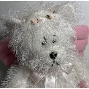 Op de afbeelding: Een witte teddybeer met een pluizige textuur, versierd met een roze strik en kleine roze rozenaccenten op het hoofd. De beer heeft zwarte ogen en een zwart gestikte neus, met roze vleugels op de achtergrond.