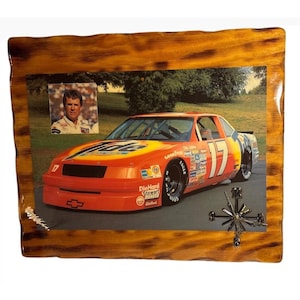 May include: A rectangular wall clock with a burnt wood frame featuring a photograph of a bright orange Tide-sponsored race car with the number 17. A small inset photo shows a man in a racing suit. The clock hands are positioned on the lower right.