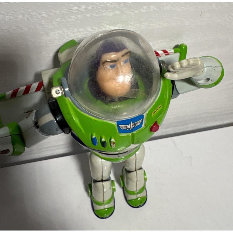 May include: A Buzz Lightyear action figure, featuring a green and white spacesuit with a clear helmet. The figure has a determined expression and outstretched arm, ready for adventure. The wings have red and white stripes.