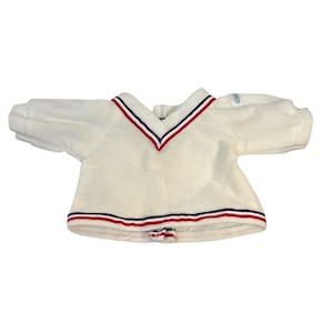 May include: Cream-colored V-neck top with red and navy blue stripes on the collar and hem. The short-sleeved shirt has a soft, textured appearance. The top is likely made of a plush material.