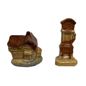 May include: Two ceramic ornaments: a miniature house with "JACK MOXX" text and a brown roof, and a brown grandfather clock. The clock has a detailed design and the word "WADE" on the base. Both are on a white background.