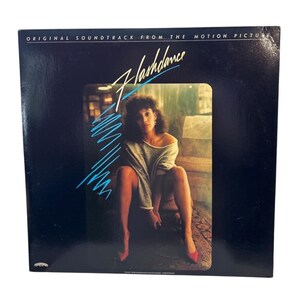 May include: Album cover for the "Flashdance" soundtrack. The cover features a woman in a gray sweater and red heels. The title is in a stylized font with blue lines. The text "ORIGINAL SOUNDTRACK FROM THE MOTION PICTURE" is at the top.