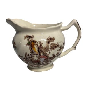 Op de afbeelding: Crème-kleurige keramische creamer met een bruin transferware landschapsontwerp. De creamer heeft een afgeronde body, een tuit en een handvat. De landschapsscène omvat gebouwen, bomen en een toren. Een klassiek stuk voor het serveren van room of melk.