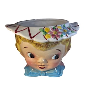 May include: A vintage ceramic planter in the shape of a child's face. The planter has blonde hair, blue eyes, and a blue bow tie. A white brim with red accents and floral details encircles the top opening. The planter is painted with bright colours.