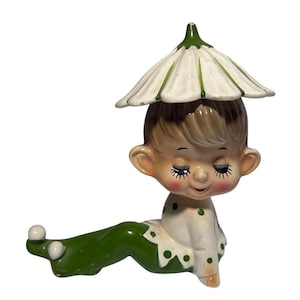 May include: A ceramic figurine of a child with closed eyes, wearing a white shirt with green polka dots and green pants. The child has a flower-shaped hat with green accents. The figurine is in a reclining position.