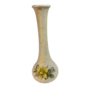 Può includere: Un vaso alto e sottile con finitura bianco sporco macchiettata. Il vaso ha una base bulbosa e un collo lungo e curvo. Un motivo floreale con fiori gialli e foglie verdi adorna la base. Il vaso è alto circa 25 cm.