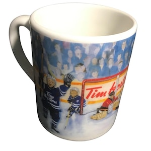 May include: White ceramic mug with a hockey scene, featuring players in blue and a goalie in red. The mug has a curved handle and the Tim Hortons logo is visible on the side.